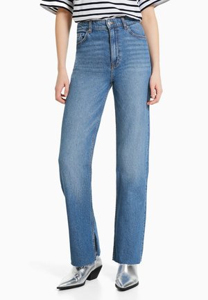 Mujer con jeans azules de pierna recta y cintura alta, con aberturas frontales y dobladillos deshilachados, combinados con botas plateadas de punta puntiaguda.