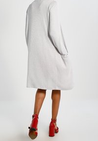 Světle šedé šaty s dlouhým rukávem, rovného střihu a bočními kapsami, doplněné červenými sandály na podpatku s otevřenou špičkou. Hladká textura.