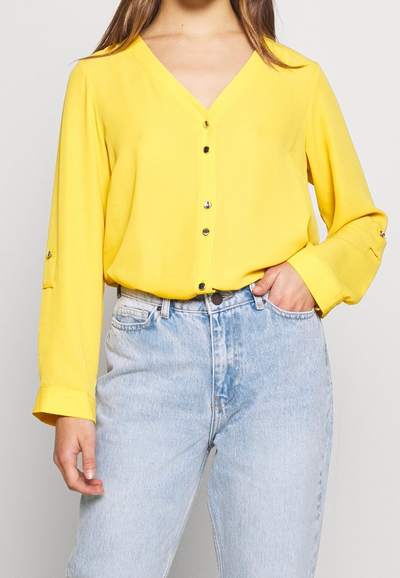 Chemise jaune à col en V avec manches longues, fermeture à boutons et détail retroussable, associée à un jean taille haute bleu clair. Texture lisse.