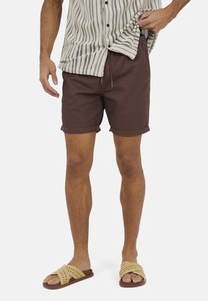 Mężczyzna w brązowych spodenkach z podwiniętym brzegiem, beżowej koszuli w paski na guziki oraz beżowych plecionych sandałach z odkrytymi palcami stoi na białym tle.