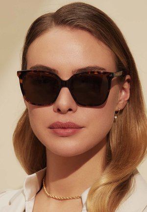 Lunettes de soleil en écaille de tortue avec des verres sombres, de forme carrée et avec des accents dorés. Complétées par un collier en chaîne dorée et une blouse légère.