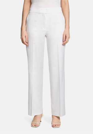 Femme portant un pantalon blanc taille haute à jambes larges associé à des sandales à talons beiges à bout ouvert, debout devant un fond uni.