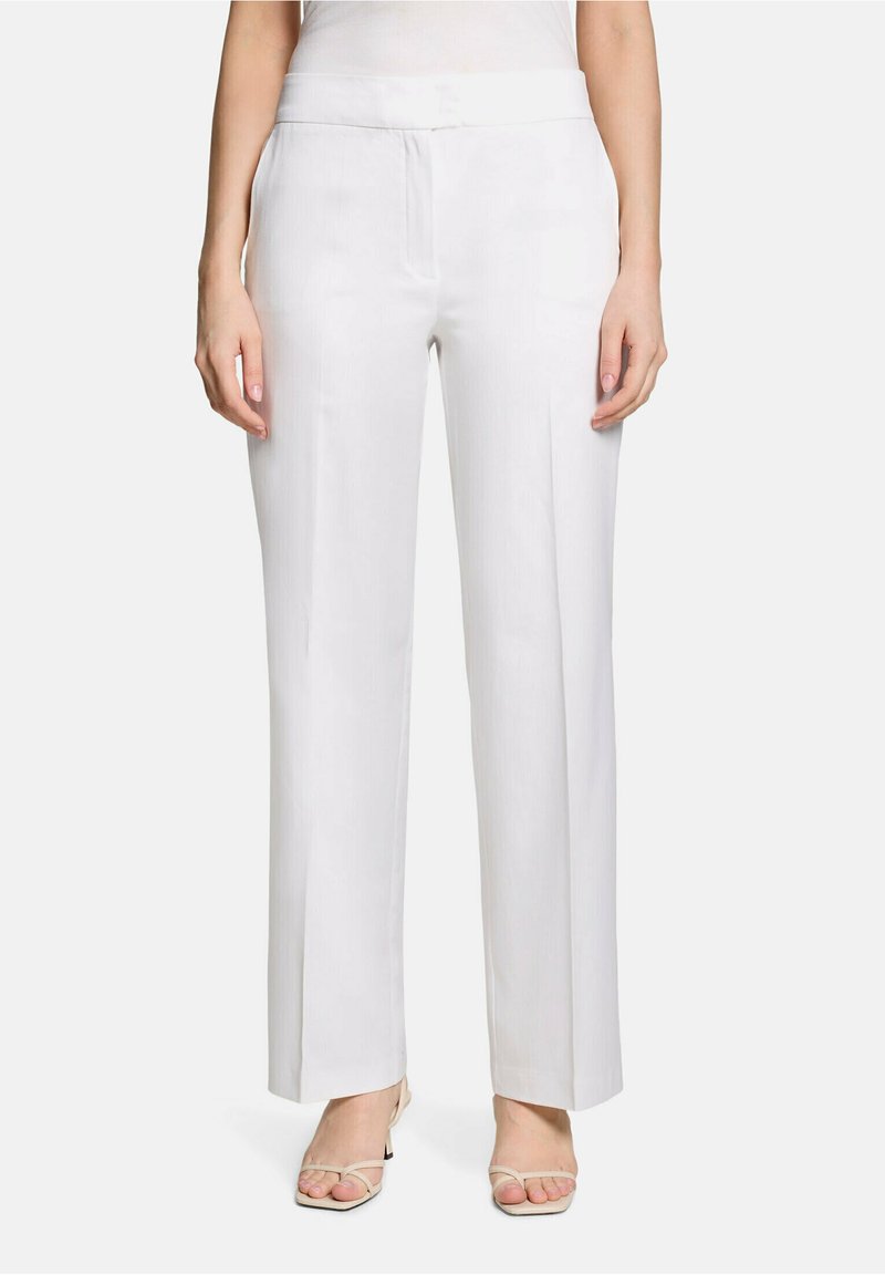 Femme portant un pantalon blanc taille haute à jambes larges associé à des sandales à talons beiges à bout ouvert, debout devant un fond uni.