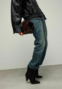 Veste aviateur en cuir noir, jeans en denim bleu délavé, pochette en cuir marron, et bottines noires cloutées avec bouts pointus et talons.