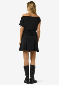 Kobieta z długimi blond włosami, ubrana w czarną bluzkę bez ramiączek, czarną spódnicę i czarne kozaki za kolano, stojąca tyłem.