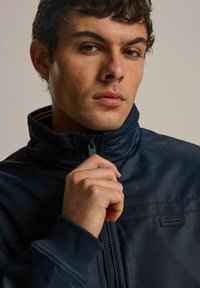 Granatowa kurtka z zamkiem na całej długości, z wysokim kołnierzem, o teksturowanej tkaninie i małym logo na piersi. Model trzyma zamek jedną ręką.