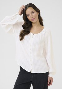 Button-up blouse in light pink, featuring a textured check pattern, square neckline, and billowy sleeves, accented by round buttons.