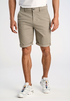 Tan, textured shorts with rolled cuffs. Front pockets and a button closure. Worn with white sneakers featuring colored accents.
