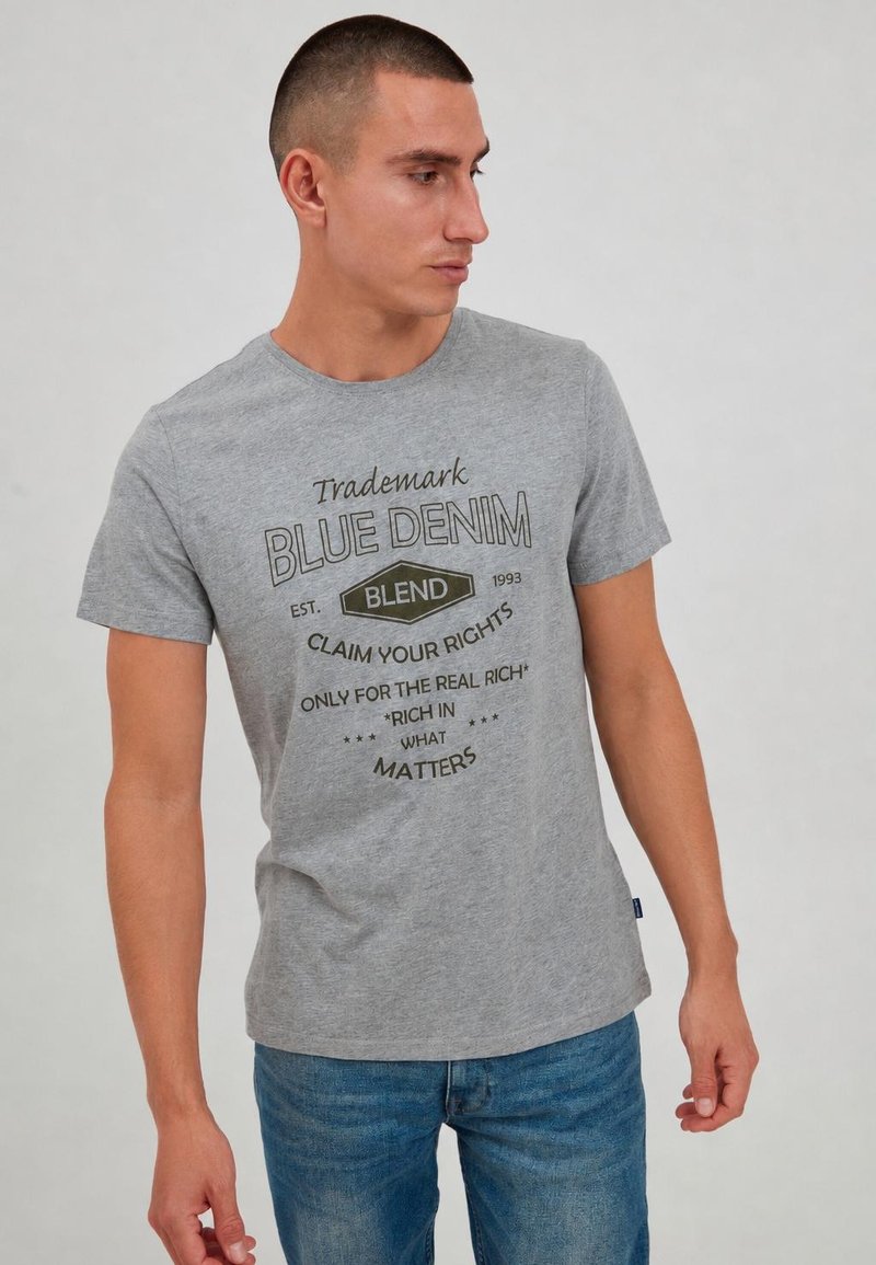 Homme portant un t-shirt graphique gris avec texte noir et un jean bleu, regardant vers sa gauche sur un fond uni.
