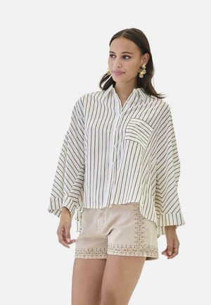 Mujer con camisa blanca de gran tamaño con finas rayas negras y pantalones cortos beige con patrones bordados, de pie contra un fondo liso.