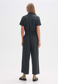 Dunkelgrüner Jumpsuit mit kurzen Ärmeln, elastischem Bund, weiten Beinen und einer glatten Textur. Kombiniert mit grünen Sandalen und einem schlichten Design.