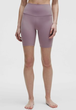 Schmal geschnittene, hoch taillierte Biker-Shorts in Mauve, getragen von einer stehenden Person mit bloßen Füßen vor einem einfachen hellen Hintergrund.