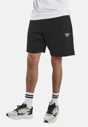 Persoon draagt zwarte Reebok shorts, witte sokken met zwarte strepen en beige-zwarte Reebok sneakers, staand tegen een effen achtergrond.