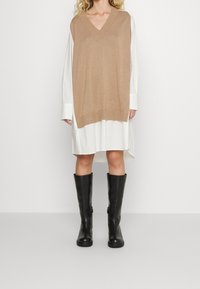 Top sans manches en tricot marron clair sur une robe chemise blanche ; le modèle porte des bottes noires jusqu'aux genoux. Détails du col en V et de l'ourlet côtelé visibles.