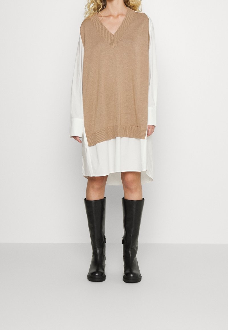 Top sans manches en tricot marron clair sur une robe chemise blanche ; le modèle porte des bottes noires jusqu'aux genoux. Détails du col en V et de l'ourlet côtelé visibles.