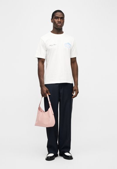 Άνδρας που φοράει λευκό γραφικό T-shirt και μπλε ναυτικό παντελόνι με ρίγες, κρατώντας μικρή ροζ τσάντα, στέκεται μπροστά σε απλό λευκό φόντο.