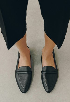 Person trägt schwarze Lederloafer und schwarze gekürzte Hosen und steht auf einem hellgrauen Boden.