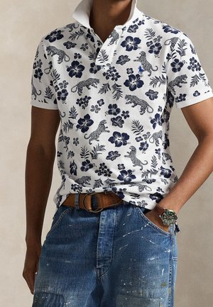 Mann trägt ein weißes Polohemd mit blauem Blumen- und Leopardenmuster, braunen Gürtel, blaue Jeans und eine Uhr mit grünem Zifferblatt am Handgelenk.