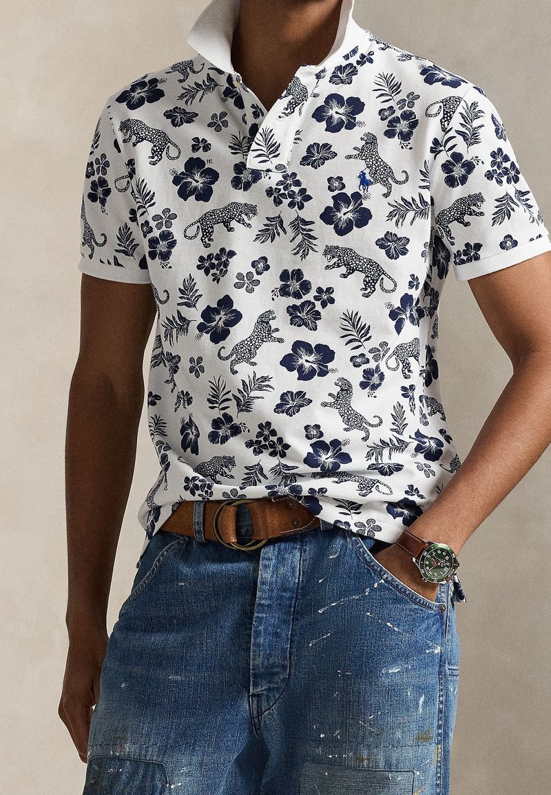 Homme portant un polo blanc avec un imprimé floral bleu et léopard, ceinture marron, jean bleu et une montre avec un cadran vert au poignet.