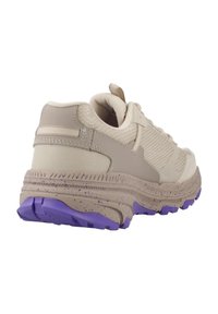 Zapatilla deportiva beige con parte superior de malla y tejido, suela texturizada y acento morado en la suela exterior. Silueta gruesa con talón reforzado.