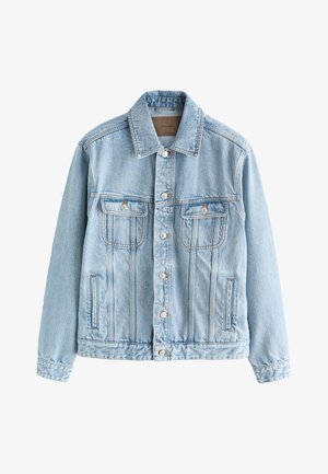 Ein junger Mann steht draußen neben einem Gebäude und trägt eine hellblaue Jeansjacke, ein gestreiftes Hemd, graue Hosen und schwarze Slipper.