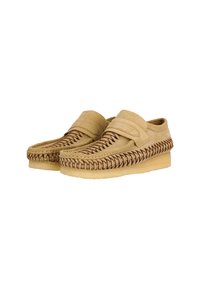 Suedeslippers i ljusbrunt med bruna flätade detaljer längs kanterna, med en platt sula och ett band över fotryggen.