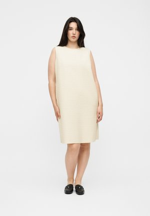 Kvinde stående iført en ærmeløs knælængde cremefarvet tekstureret kjole og sorte slip-on sko mod en ensfarvet lys baggrund.