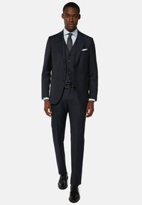 Granatowy garnitur trzyczęściowy z dopasowaną marynarką, kamizelką i zwężanymi spodniami, wykonany z gładkiej tkaniny, zestawiony z czarnymi butami i jasnoniebieską koszulą.