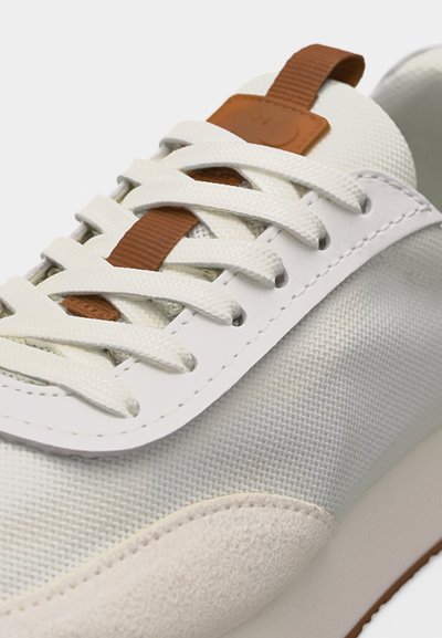Baskets blanches avec tissu texturé, panneau latéral lisse, lacets blancs et languette marron sur la languette.