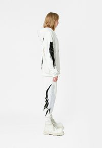 Sudadera blanca con capucha y pantalones deportivos con gráficos laterales de alas en blanco y negro; combinados con botas blancas gruesas; tejido suave y corte relajado.