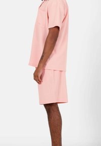 Camicia rosa chiaro a maniche corte con tasca e pantaloni corti abbinati. Entrambi presentano un tessuto texturizzato. I pantaloni corti hanno un logo del marchio discreto.