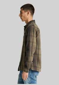 Jonge man in een bruin en zwart geruit shirt en blauwe spijkerbroek, staand in profiel tegen een effen grijze achtergrond.