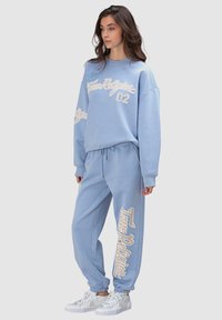 Sweat-shirt bleu clair et joggers assortis en tissu doux, présentant de grands textes et logos blancs imprimés. Stylés avec des poignets élastiques et un cordon de serrage.