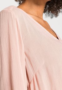 Blusa de color rosa claro con escote en V, hecha de tejido texturizado. Presenta mangas largas y un corte holgado con pliegues verticales.