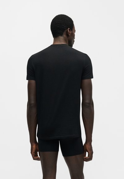 Άνδρας με κοντά σγουρά μαλλιά, που φοράει απλό μαύρο κοντομάνικο μπλουζάκι και μαύρα σορτς, στέκεται με την πλάτη προς τα εμπρός σε λευκό φόντο.