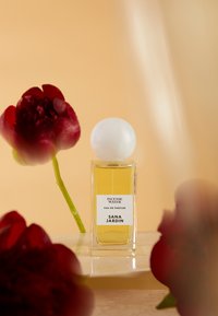 Bouteille de parfum en verre transparent contenant un liquide jaune, avec un bouchon sphérique blanc et une étiquette indiquant "EAU D'ENCENS" et "SANA JARDIN", entourée de fleurs rouges.