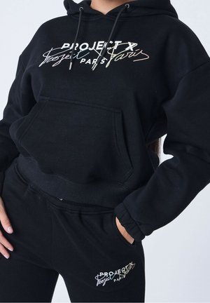 Ensemble sweat à capuche noir et pantalon de survêtement avec le logo "PROJECT X Paris", mains dans les poches, recadré à mi-torse montrant une tenue sportive décontractée.