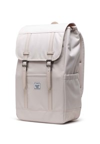 Sac à dos en tissu beige avec un rabat, deux sangles avant, des coutures détaillées, un panneau arrière en maille et une petite étiquette logo à l'avant.
