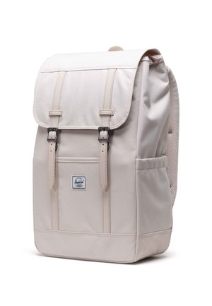 Beige stoffen rugzak met een flap, twee voorste banden, gedetailleerde stiksels, een mesh rugpaneel en een klein logo-label aan de voorkant.