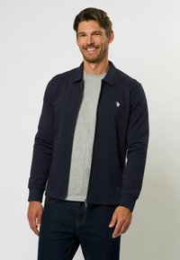 Jūras spēku zip-up jaka ar apkakli, ar krūšu logotipu, uzvilkta pāri pelēkai krekla. Materiāls izskatās gluds un ar pieskaņotu dizainu.