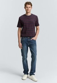 Triibuline mereväe lühikeste varrukatega särk punaste detailidega, koos siniste teksadega. Modell kannab valgeid tenniseid ja seisab heleda tausta ees.