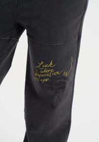 Svarta bomullsbukser med gul broderad text och en blekt tiger grafik på benet. Designen har sydda detaljer och en avslappnad passform.