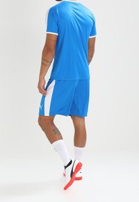 Blå idrottströja och shorts med vita accenter, tillverkade av lättviktsmaterial. Har logotyp på shortsen, bärs med vita strumpor och svarta skor.