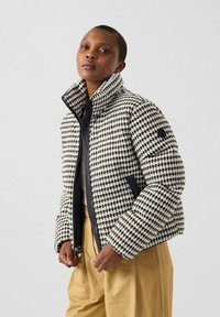 Vrouw in een zwart-wit houndstooth patroon pufferjack, beige broek, staande tegen een effen lichtgrijze achtergrond.