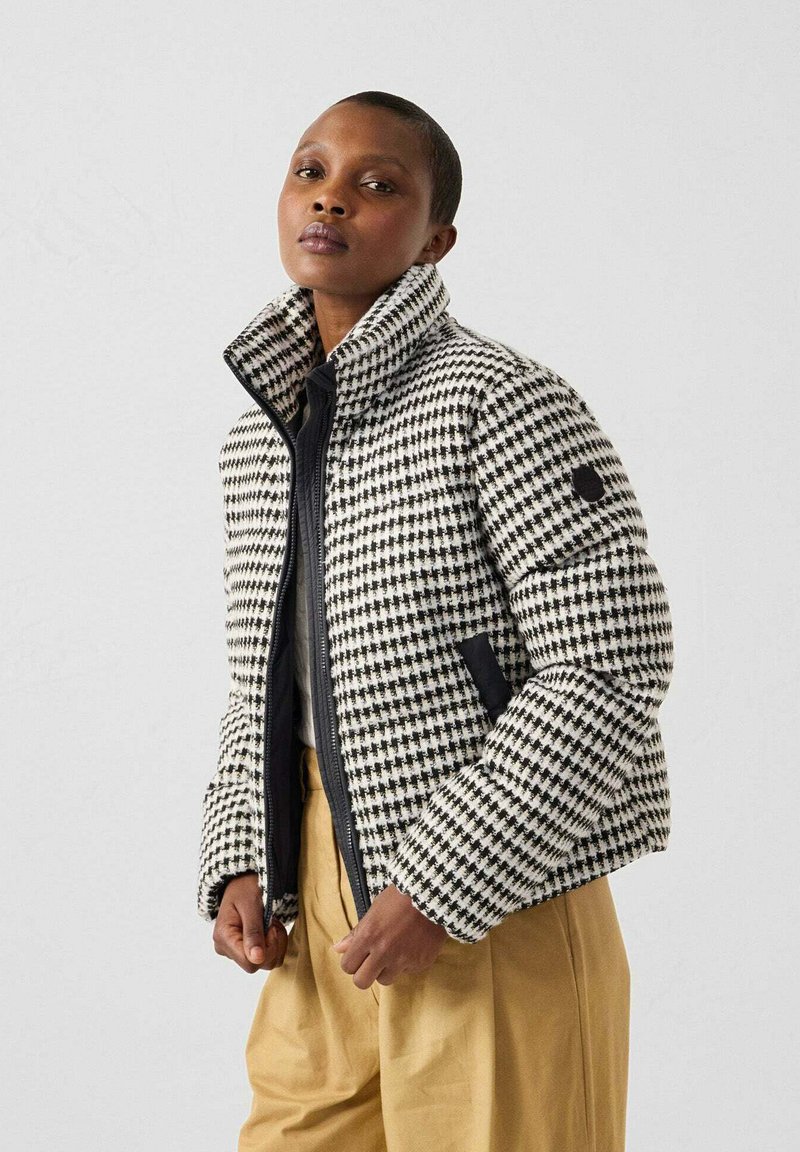 Vrouw in een zwart-wit houndstooth patroon pufferjack, beige broek, staande tegen een effen lichtgrijze achtergrond.