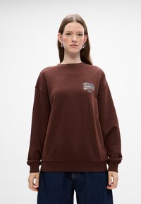 Sudadera marrón con cuello redondo, hombros caídos y logotipo bordado de "Tommy". Presenta puños y dobladillo acanalados, con una textura de algodón suave.