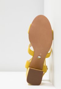 Sandale en daim jaune avec talon carré, semelle tournée vers le haut, taille 38 indiquée sur la semelle, sur fond blanc.