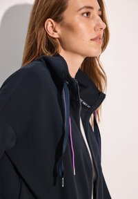 Giacca navy con zip e colletto alto, caratterizzata da lacci a contrasto blu e viola. Tessuto morbido con una consistenza liscia.