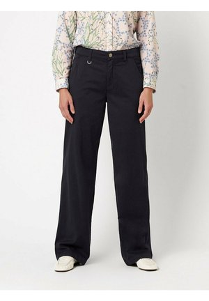 Kobieta w czarnych szerokich spodniach, kwiecistej bluzce z długim rękawem i białych wsuwanych butach, stojąca na jednolitym tle.