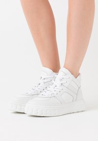 Bílé vysoké tenisky s tlustými podrážkami a tkaničkami nošené na holých nohách na jednoduchém bílém pozadí.